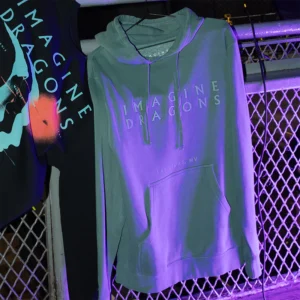 Imagine Dragons merch pullover hoodie in teal featuring embossed text design with Imagine Dragons and Las Vegas NV on the front Displayed under purple lighting alongside another band themed sweatshirt