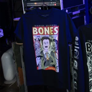 Imagine Dragons merch - A black T-shirt featuring a graphic comic-style design inspired by the song "Bones." The print includes a central character in a suit with a shocked expression, surrounded by zombies. The word "BONES" is prominently displayed at the top in bold red letters, with "IMAGINE DRAGONS" written above it. The dark setting and stage equipment in the background suggest a concert or backstage environment.