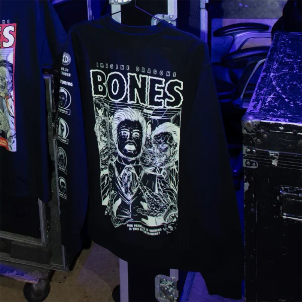 Black Glow in the Dark Bones album merch crewneck sweatshirt hanging on a rack in a dimly lit backstage setting The design features eerie horror inspired graphics bold BONES text and Imagine Dragons branding The sleeve has circular logos and the glow effect is subtly visible under the lighting